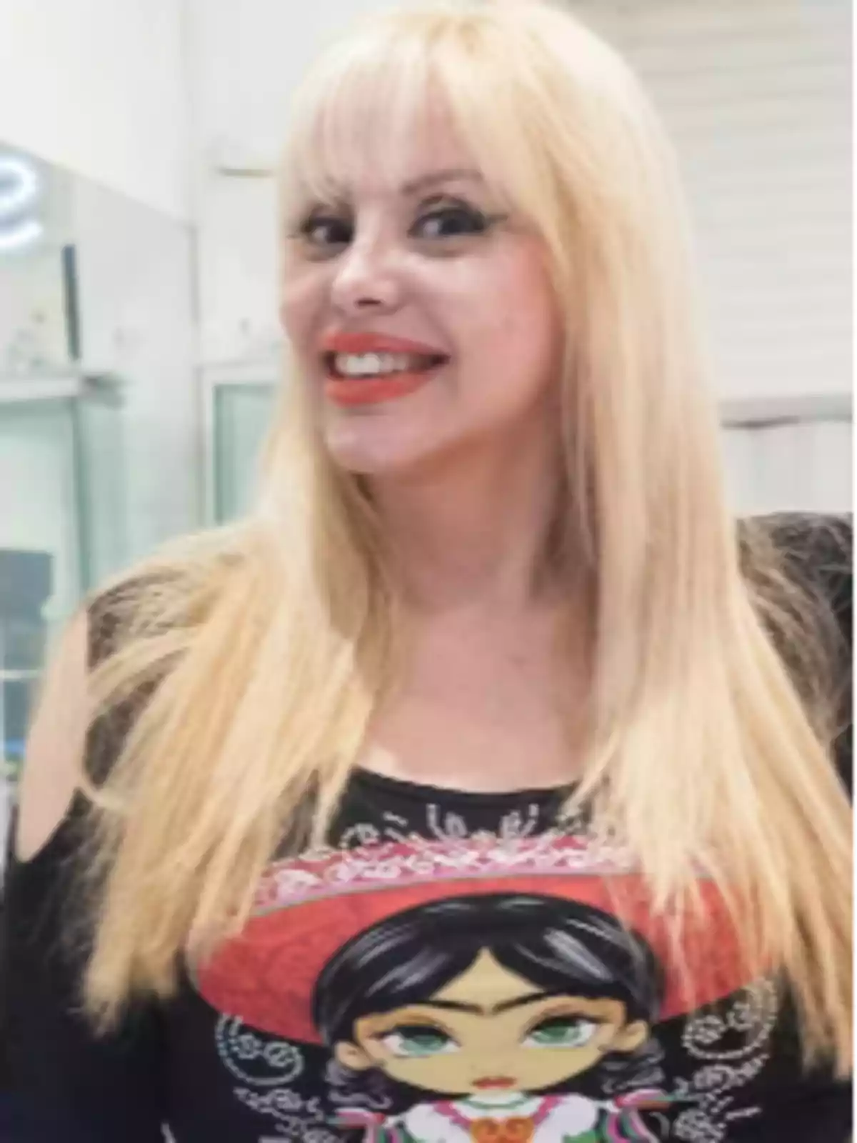 Una persona con cabello rubio y una camiseta con un diseño colorido sonríe frente a la cámara en un entorno interior. Una persona con cabello rubio y una camiseta con un diseño colorido sonríe frente a la cámara en un entorno interior.
