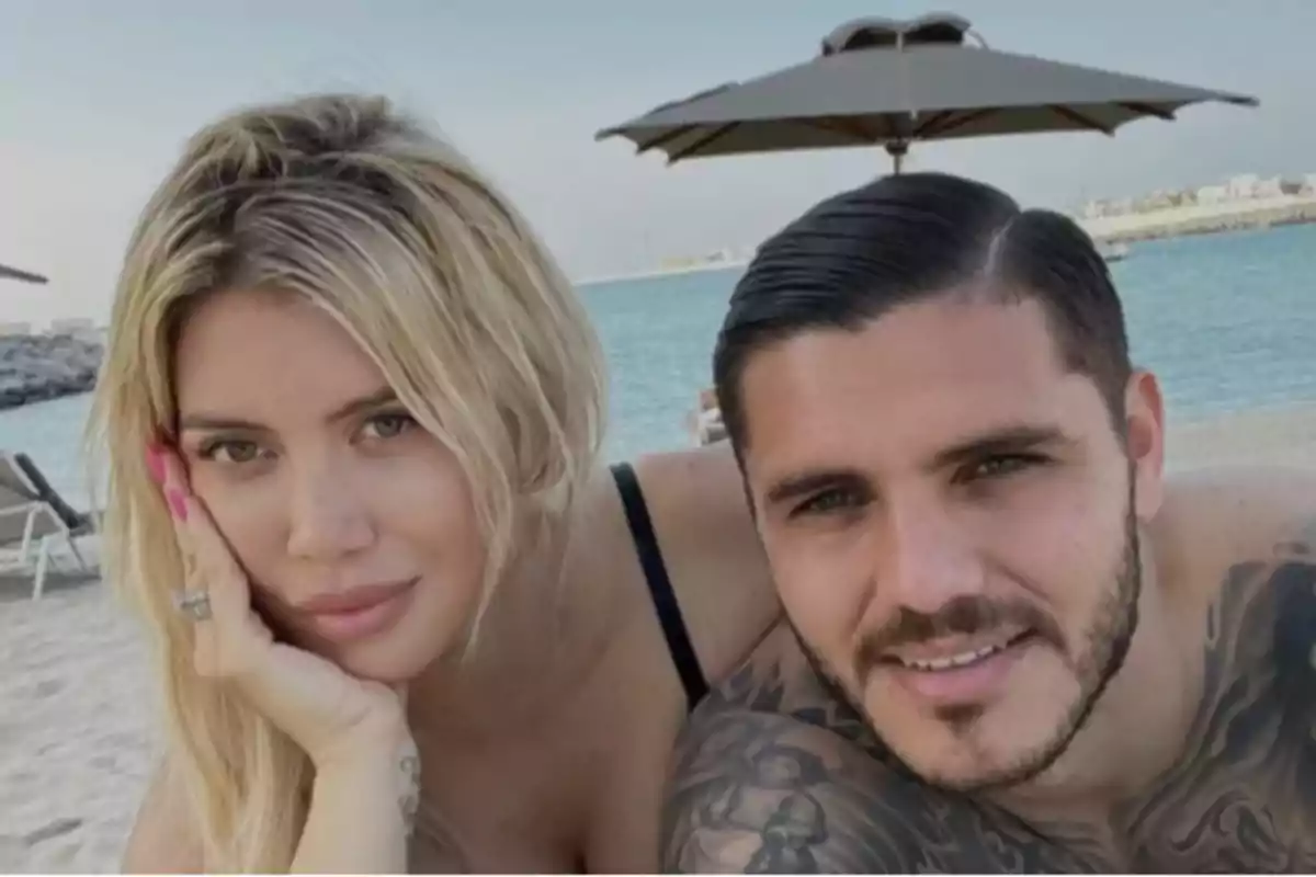A couple smiles while on the beach with the sea and an umbrella in the background.