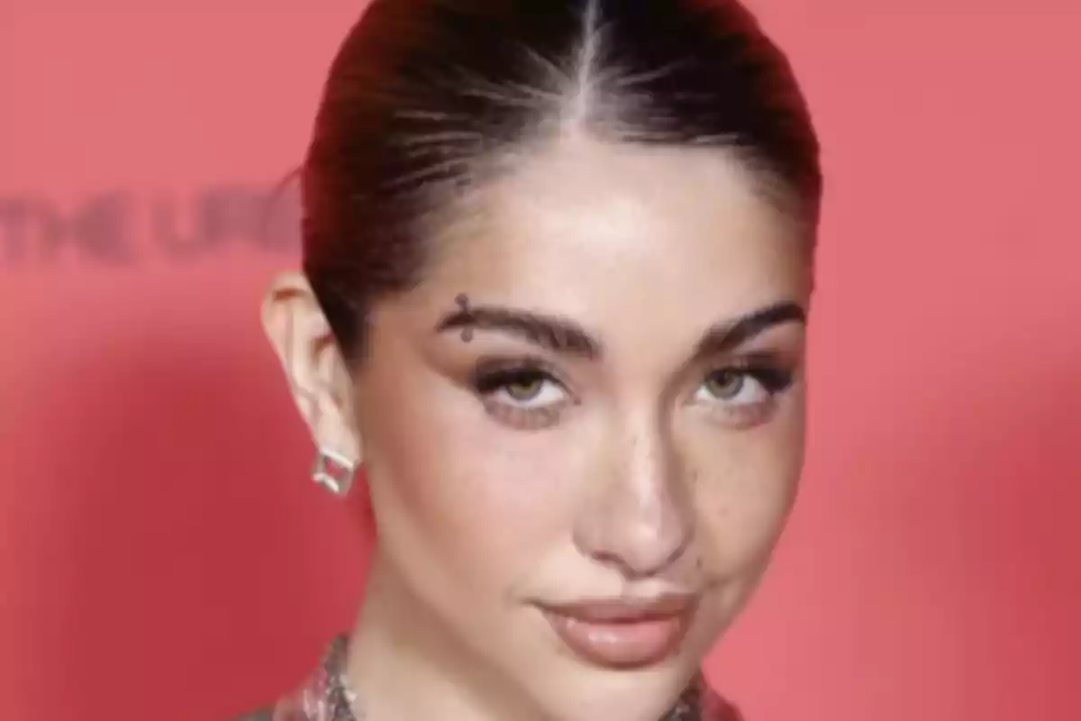 Una persona con cabello recogido y maquillaje sutil posa frente a un fondo rojo.