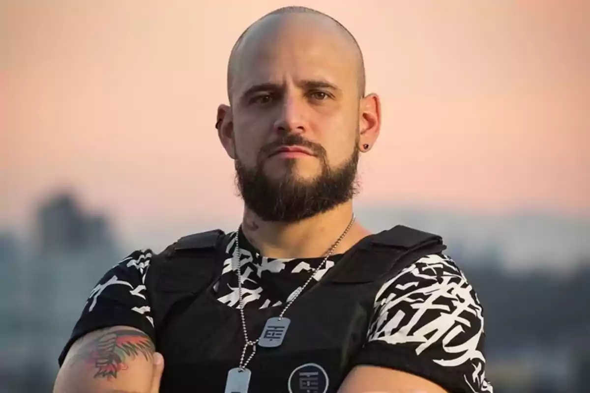 A man with a beard and a shaved head wears a graphic design T-jersey and a black vest while staring intently at the camera with a sunset sky in the background.