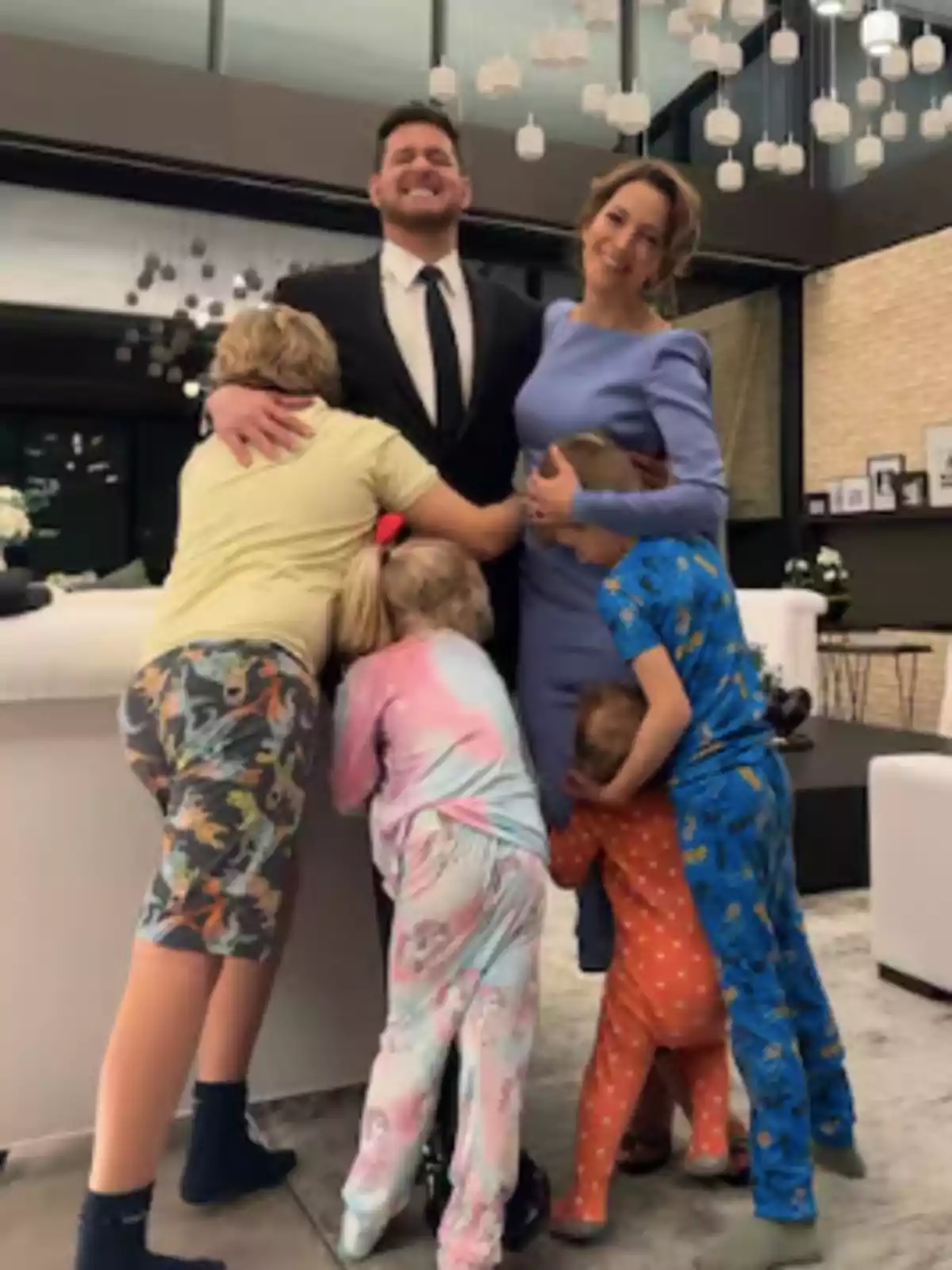 A smiling couple is surrounded by four children in colorful pajamas in a modern living room with hanging lights. A smiling couple is surrounded by four children in colorful pajamas in a modern living room with hanging lights.