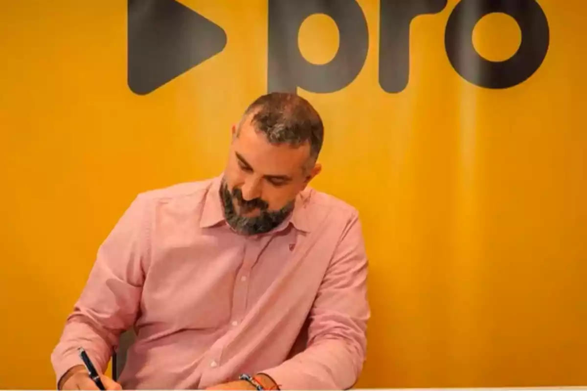 A man with a beard and a pink jersey writes on a paper in front of a yellow background with black letters.