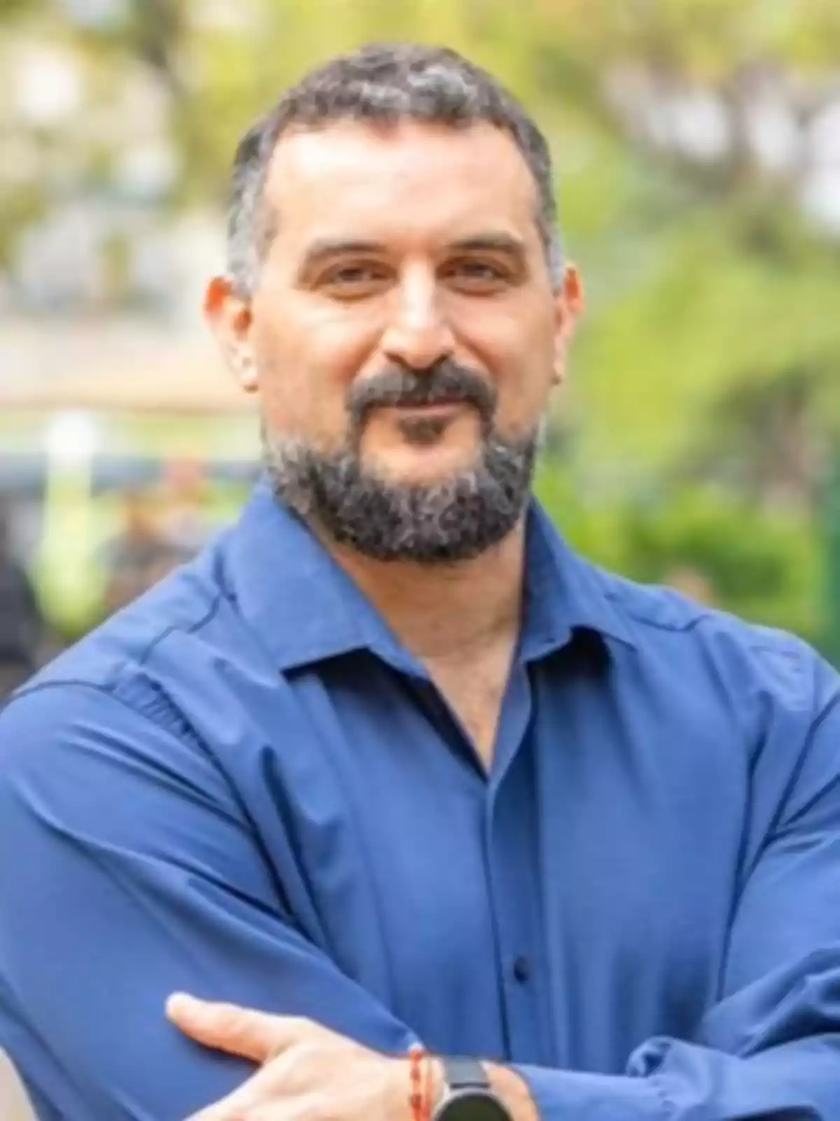 A man with a beard and short hair, wearing a blue jersey, is standing outdoors with his arms crossed and a blurred background of vegetation. A man with a beard and short hair, wearing a blue jersey, is standing outdoors with his arms crossed and a blurred background of vegetation.