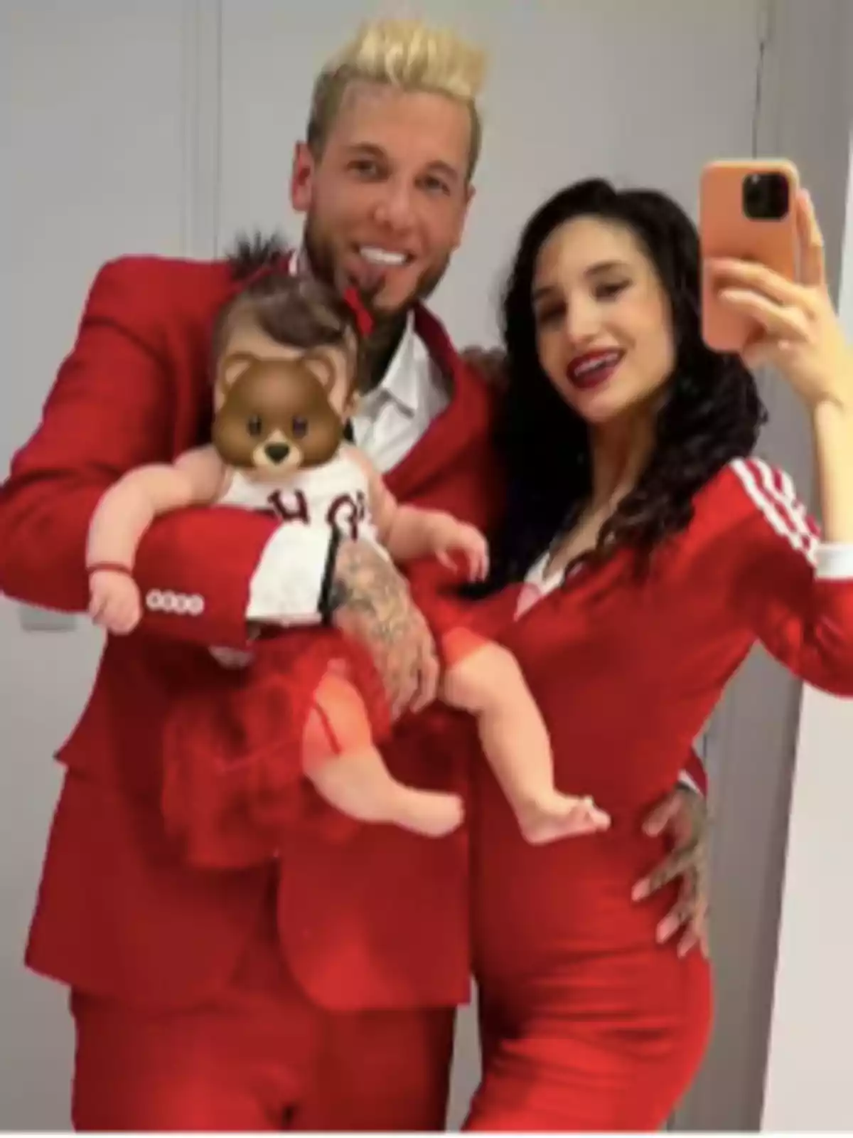 A couple dressed in red poses for a selfie in a mirror while holding a baby with a bear emoji on its face. A couple dressed in red poses for a selfie in a mirror while holding a baby with a bear emoji on its face.