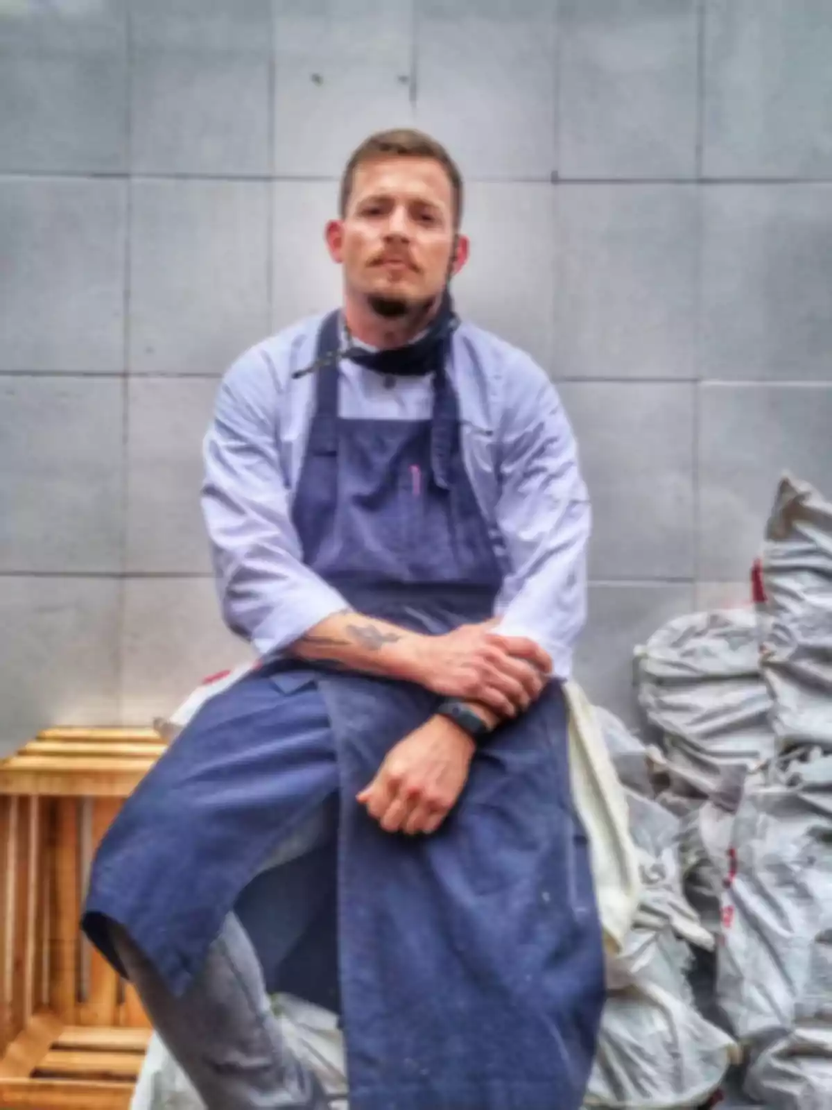 A man in a blue apron is sitting in front of a gray tiled wall, with bags stacked beside him. A man in a blue apron is sitting in front of a gray tiled wall, with bags stacked beside him.