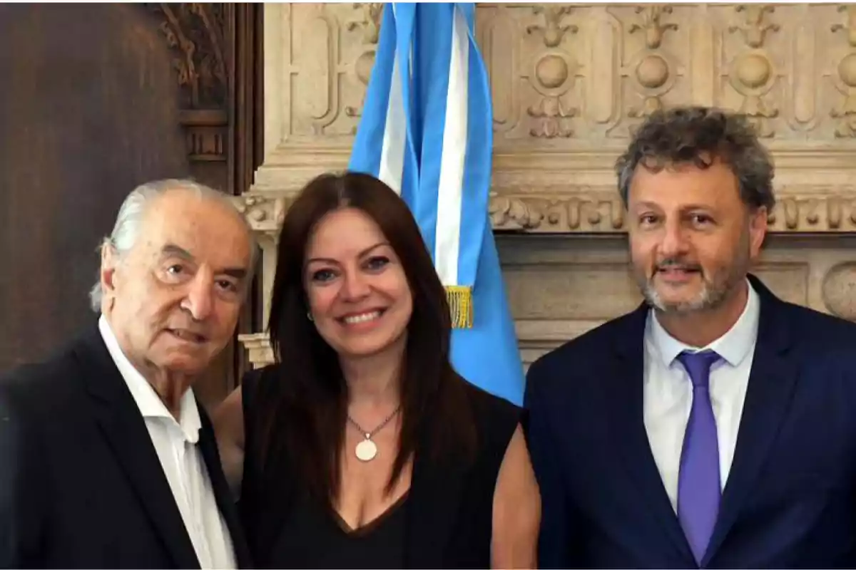 Three people pose together in front of a blue and white flag in an elegant setting. Three people pose together in front of a blue and white flag in an elegant setting.