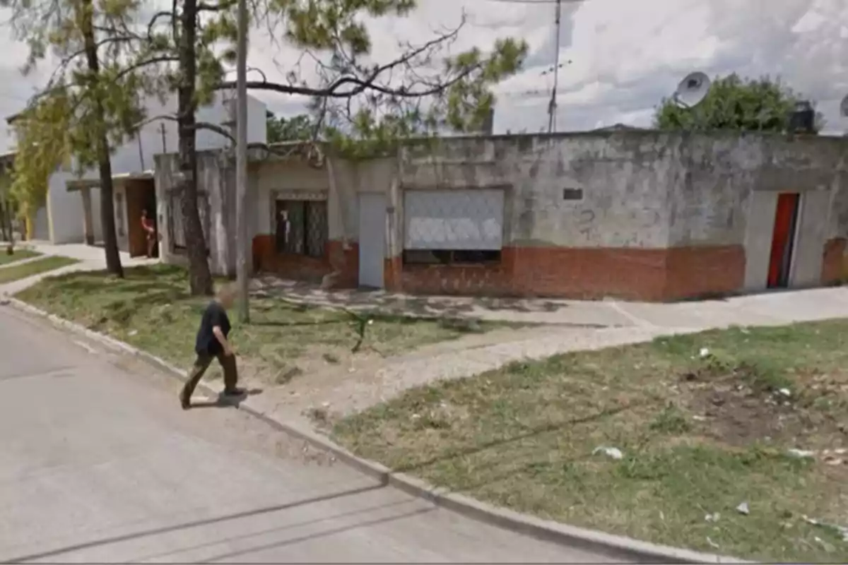 Un hombre camina por la acera frente a una casa de aspecto desgastado con paredes de ladrillo y cemento, rodeada de árboles y vegetación. Un hombre camina por la acera frente a una casa de aspecto desgastado con paredes de ladrillo y cemento, rodeada de árboles y vegetación.