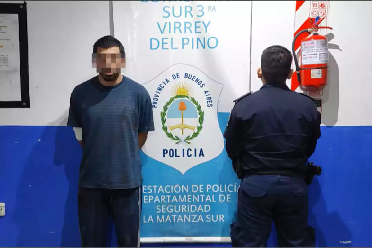 A man with a pixelated face stands next to a police officer in front of a sign for the La Matanza Sur police station in Buenos Aires.