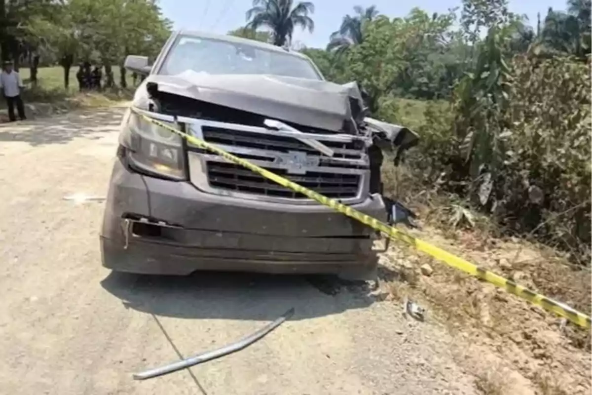 A vehicle with a damaged front is stopped on a rural road surrounded by vegetation and caution tape.