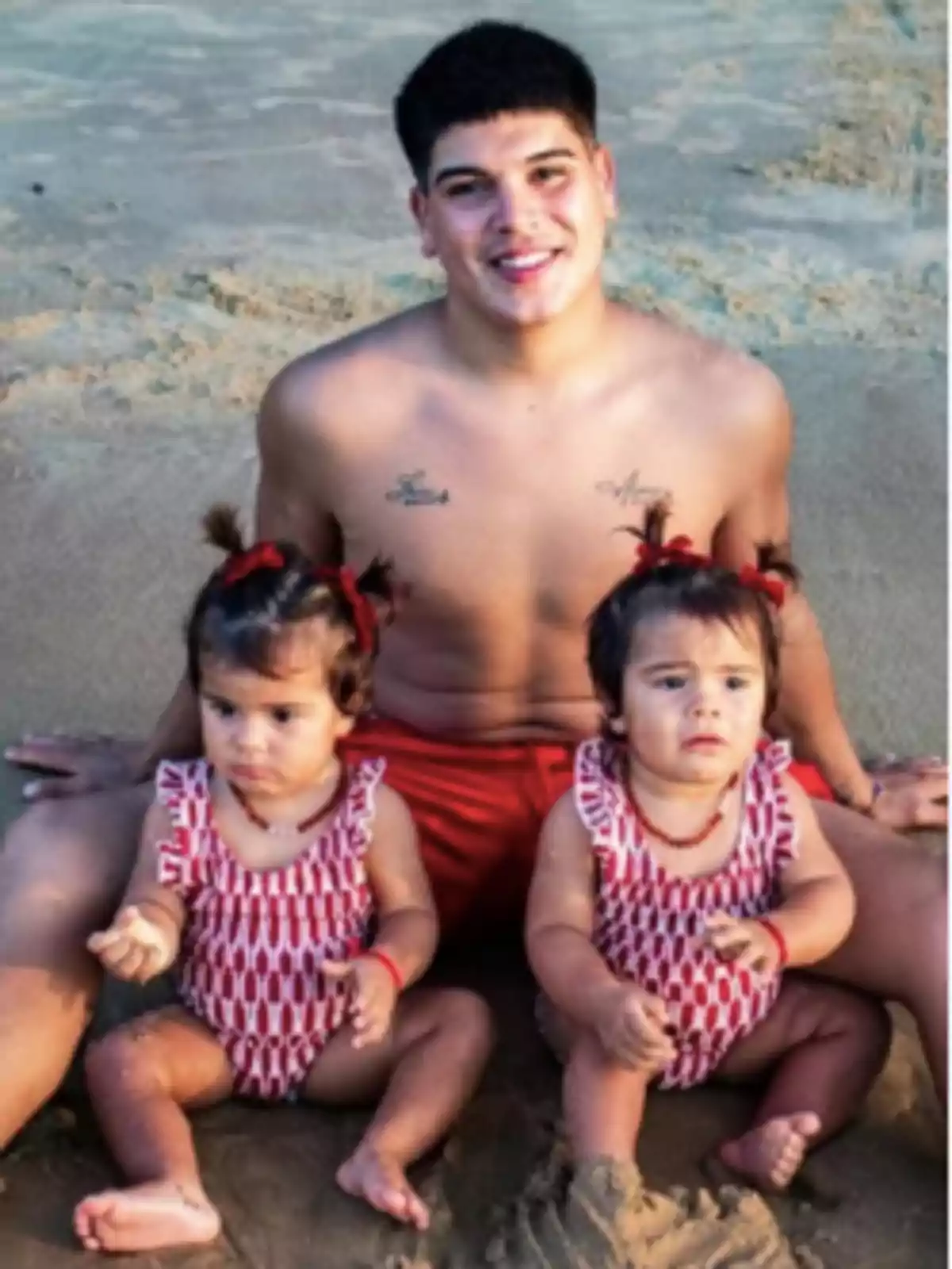 Un hombre joven sin camisa está sentado en la arena de la playa con dos niñas pequeñas, ambas con trajes de baño rojos con estampado blanco y moños rojos en el cabello. Un hombre joven sin camisa está sentado en la arena de la playa con dos niñas pequeñas, ambas con trajes de baño rojos con estampado blanco y moños rojos en el cabello.