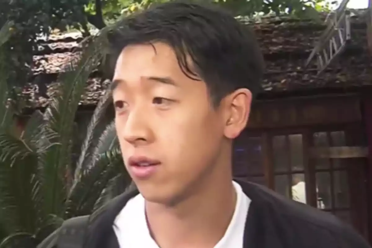 Un hombre joven con cabello oscuro y expresión seria está al aire libre, con vegetación y una estructura de madera en el fondo. Un hombre joven con cabello oscuro y expresión seria está al aire libre, con vegetación y una estructura de madera en el fondo.