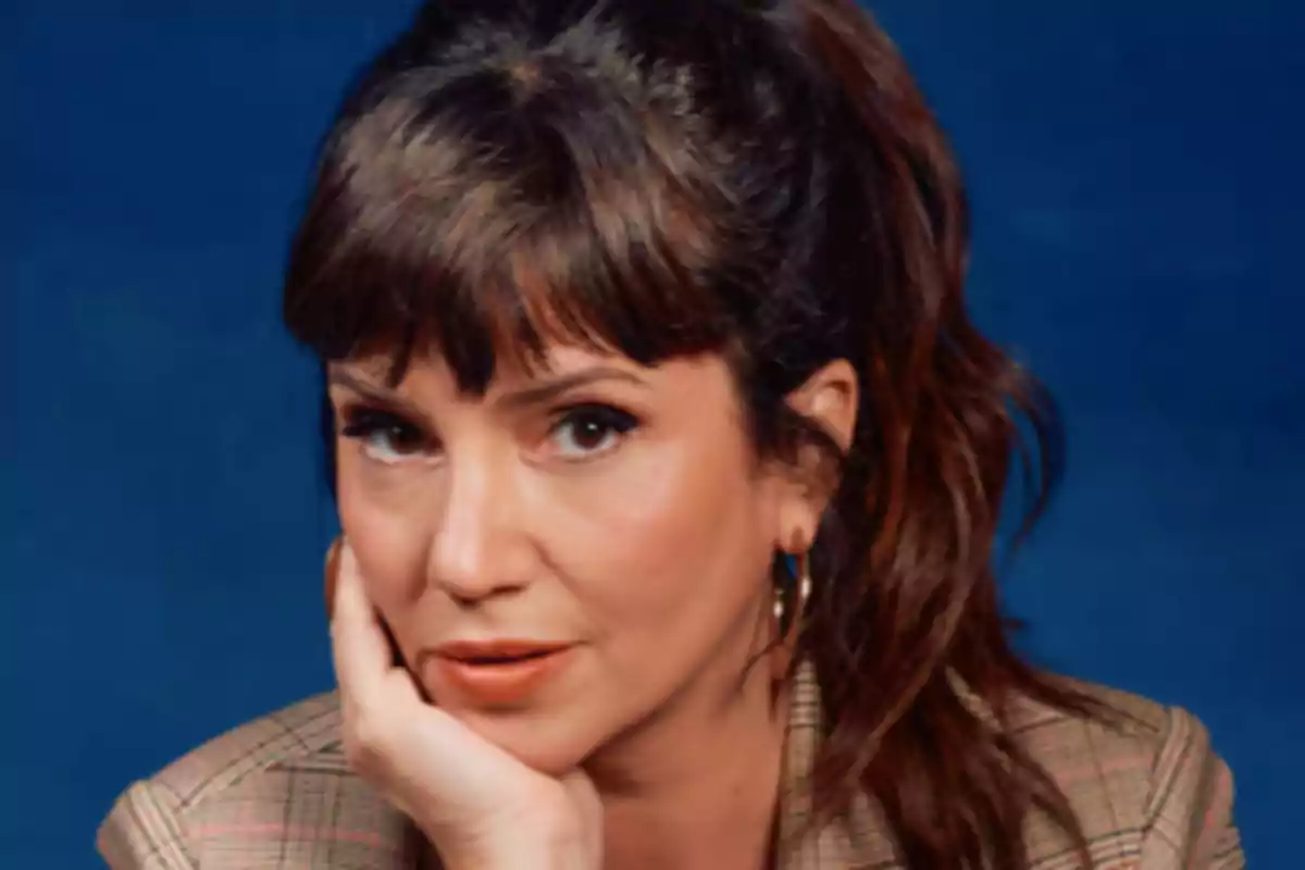 Una persona con cabello castaño y flequillo mira a la cámara con la mano apoyada en el rostro sobre un fondo azul.