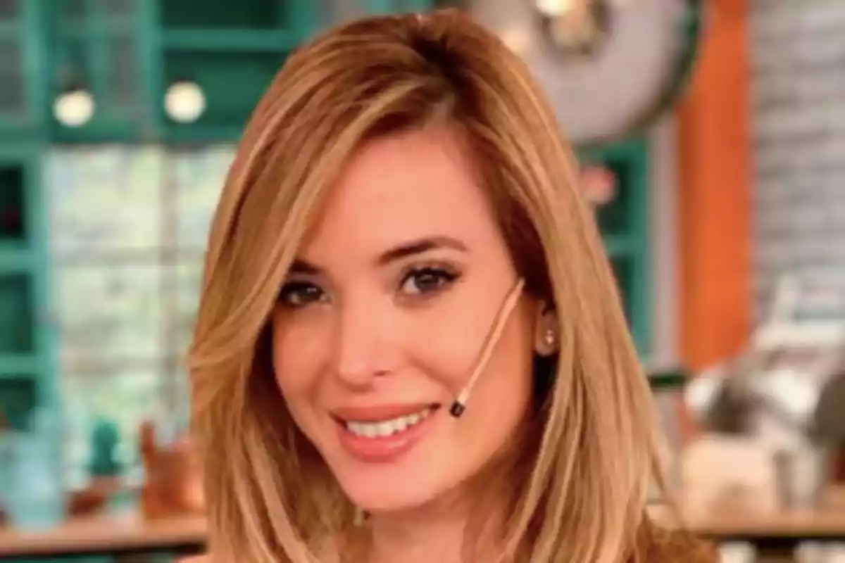 A person with blonde hair and a microphone on their cheek smiling in an indoor setting.