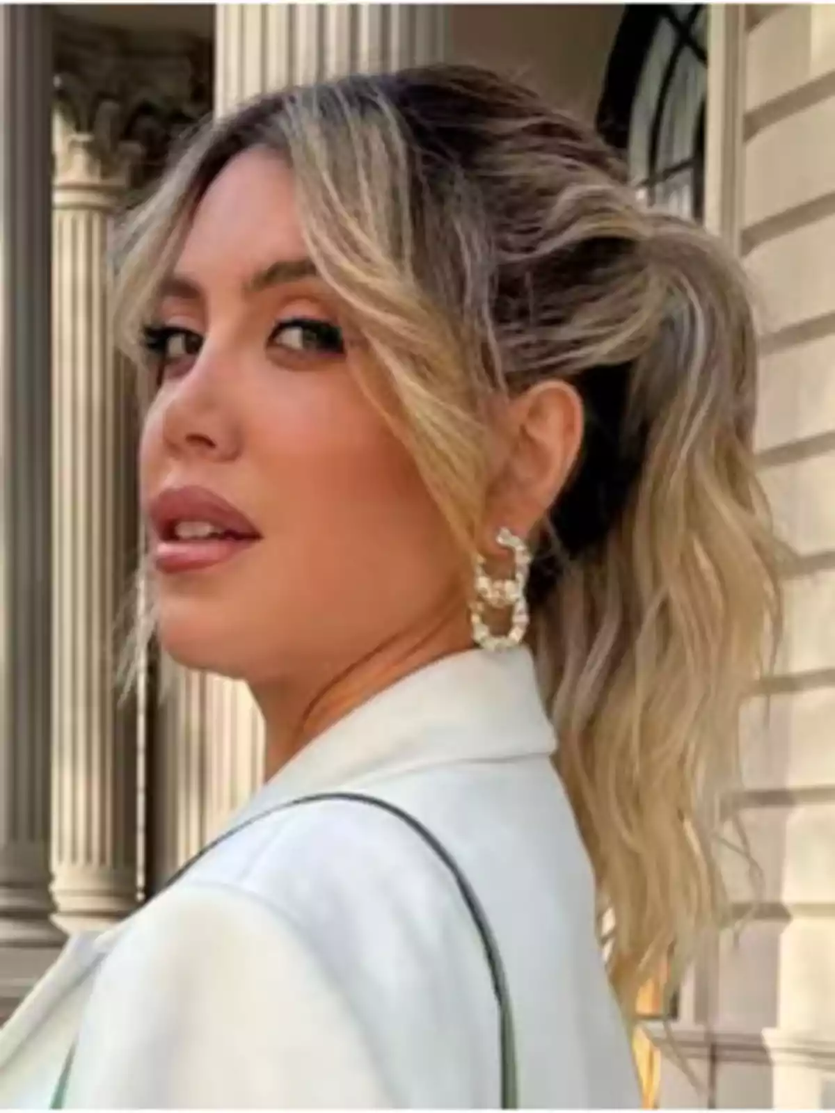 Blonde-haired woman with her hair up and large earrings, wearing a white jacket in front of a building with columns. Blonde-haired woman with her hair up and large earrings, wearing a white jacket in front of a building with columns.