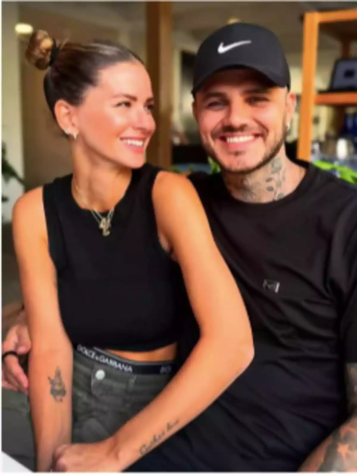 A smiling couple sitting indoors, both wearing casual clothes and having visible tattoos. A smiling couple sitting indoors, both wearing casual clothes and having visible tattoos.