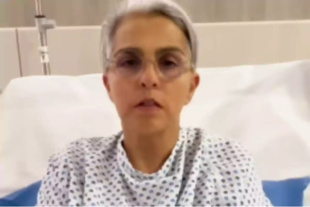 Una persona con bata de hospital y gafas está sentada en una cama de hospital.