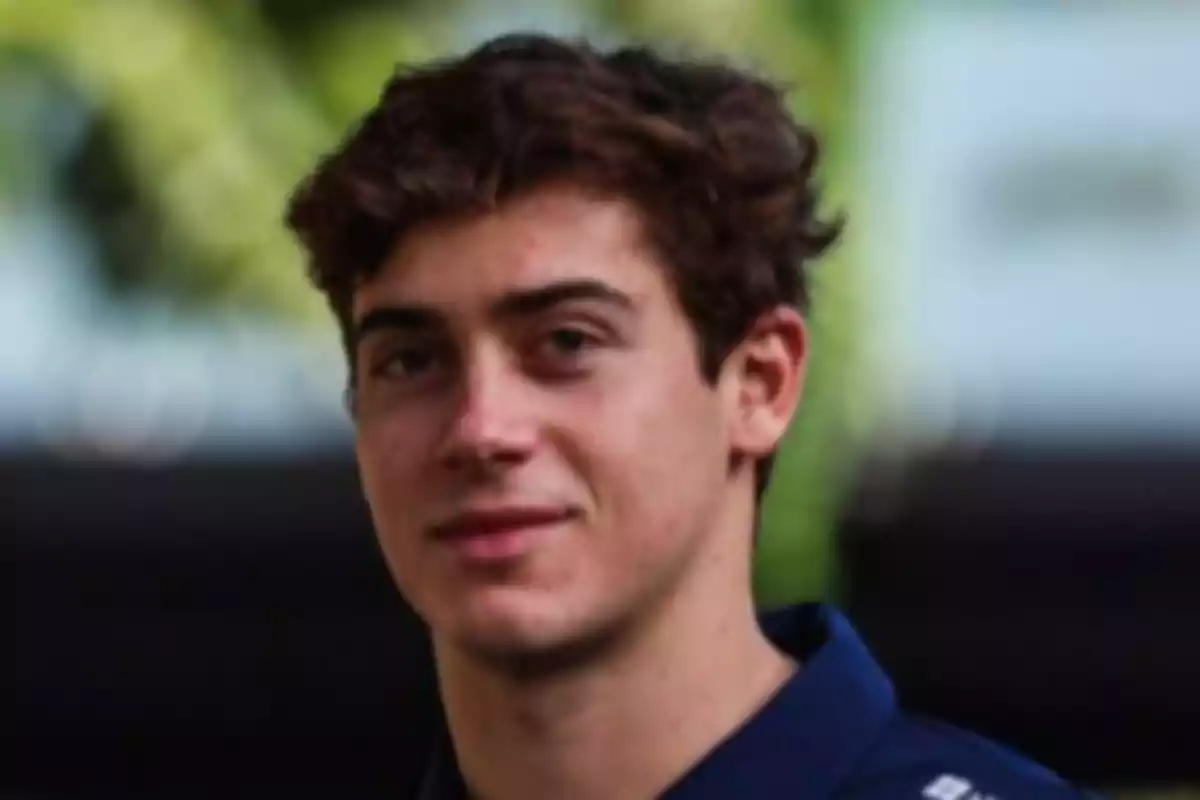 A young man with brown hair and a blue jersey slightly smiles while looking at the camera in an outdoor setting.