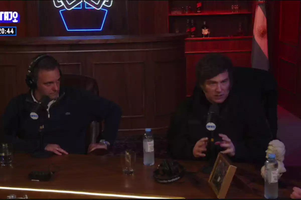 Dos personas sentadas en un estudio de grabación con micrófonos, una de ellas gesticulando mientras habla, hay una bandera de Argentina en el fondo y botellas sobre la mesa. Dos personas sentadas en un estudio de grabación con micrófonos, una de ellas gesticulando mientras habla, hay una bandera de Argentina en el fondo y botellas sobre la mesa.