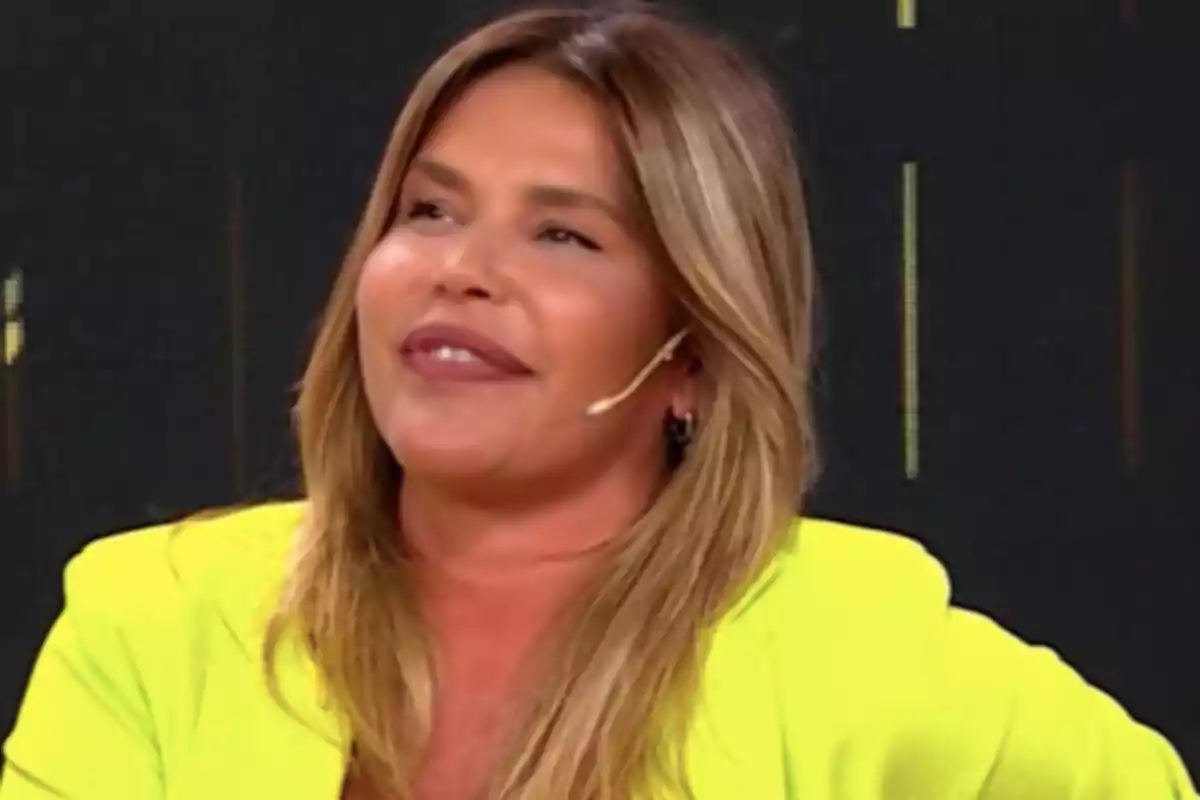 Una persona con cabello rubio y un micrófono en la oreja, vistiendo una chaqueta amarilla, sonríe mientras participa en un programa de televisión.