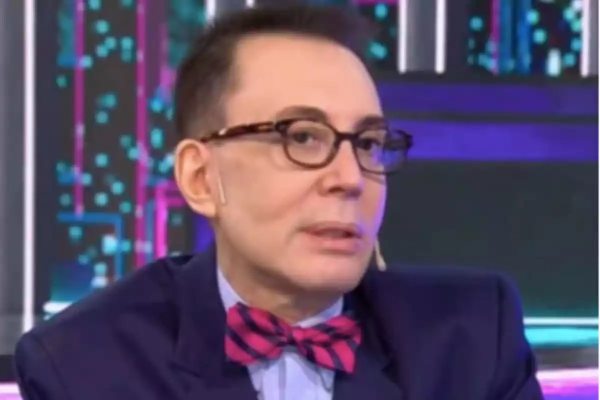 Un hombre con gafas y pajarita rosa y azul aparece en un programa de televisión con un fondo colorido.