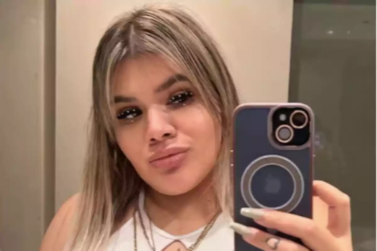 Una persona con cabello rubio y maquillaje llamativo se toma una selfie frente a un espejo usando un teléfono móvil.