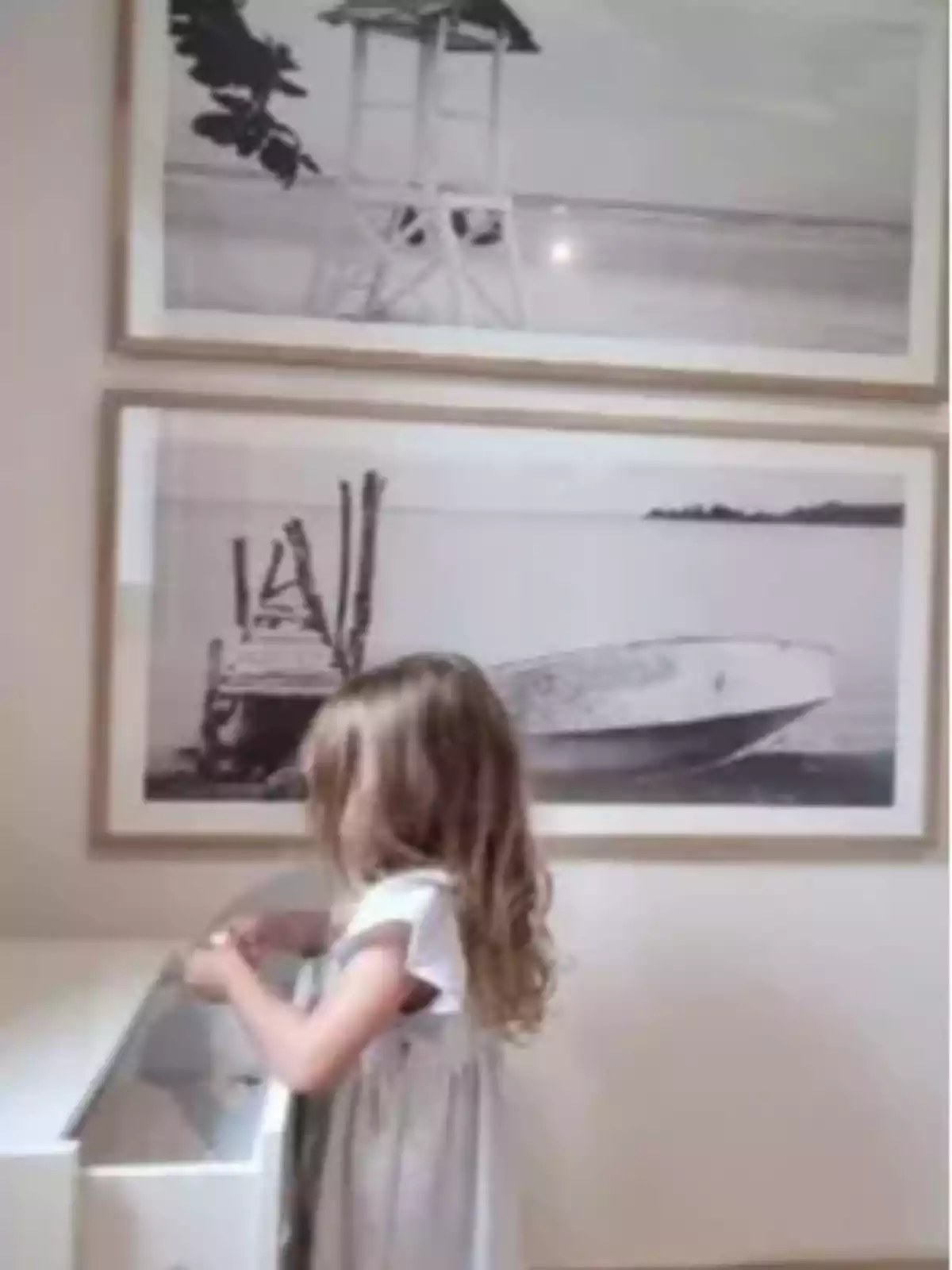 Una niña con vestido claro está de pie frente a un mueble blanco, con dos cuadros en blanco y negro de paisajes marinos colgados en la pared detrás de ella. Una niña con vestido claro está de pie frente a un mueble blanco, con dos cuadros en blanco y negro de paisajes marinos colgados en la pared detrás de ella.