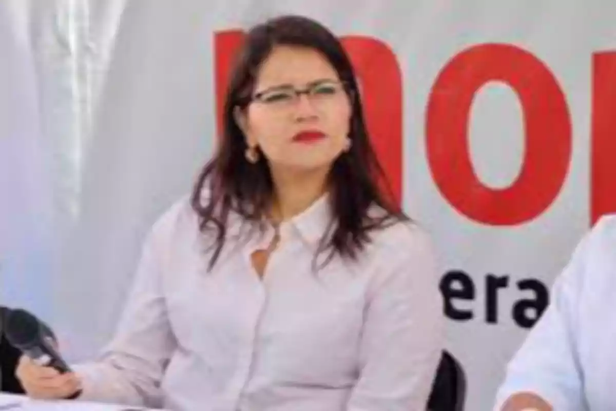 Una persona con gafas y camisa blanca sostiene un micrófono mientras está sentada frente a un fondo con letras rojas.