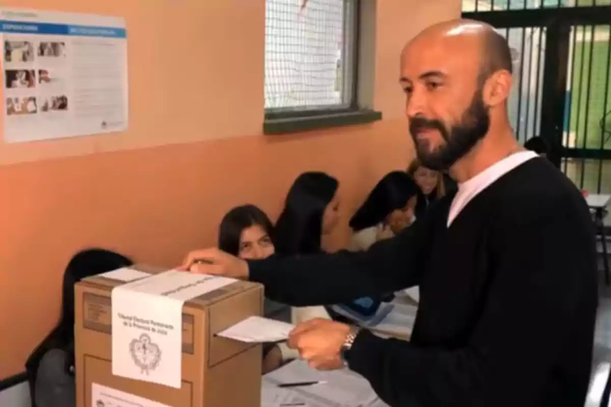 Un hombre emite su voto en una urna en un centro de votación, mientras varias personas están sentadas al fondo. Un hombre emite su voto en una urna en un centro de votación, mientras varias personas están sentadas al fondo.