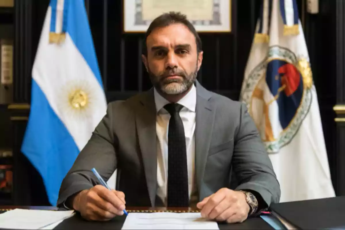 Un hombre con traje y corbata está sentado en un escritorio con banderas detrás de él. Un hombre con traje y corbata está sentado en un escritorio con banderas detrás de él.