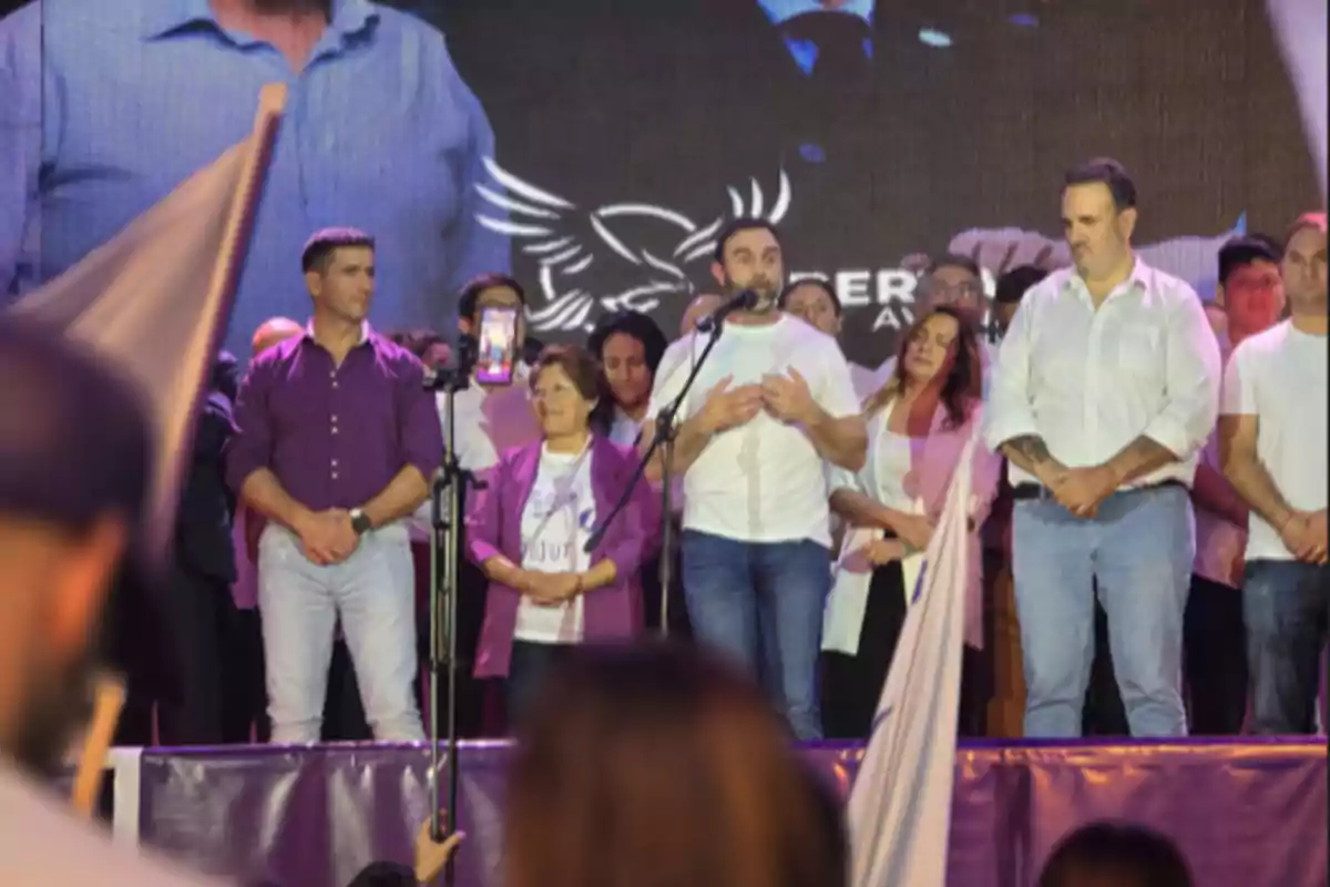 Un grupo de personas está en un escenario durante un evento, con un hombre hablando al micrófono mientras otros lo rodean, algunos de ellos visten ropa en tonos morados y blancos, y hay una pantalla grande detrás mostrando una imagen ampliada.