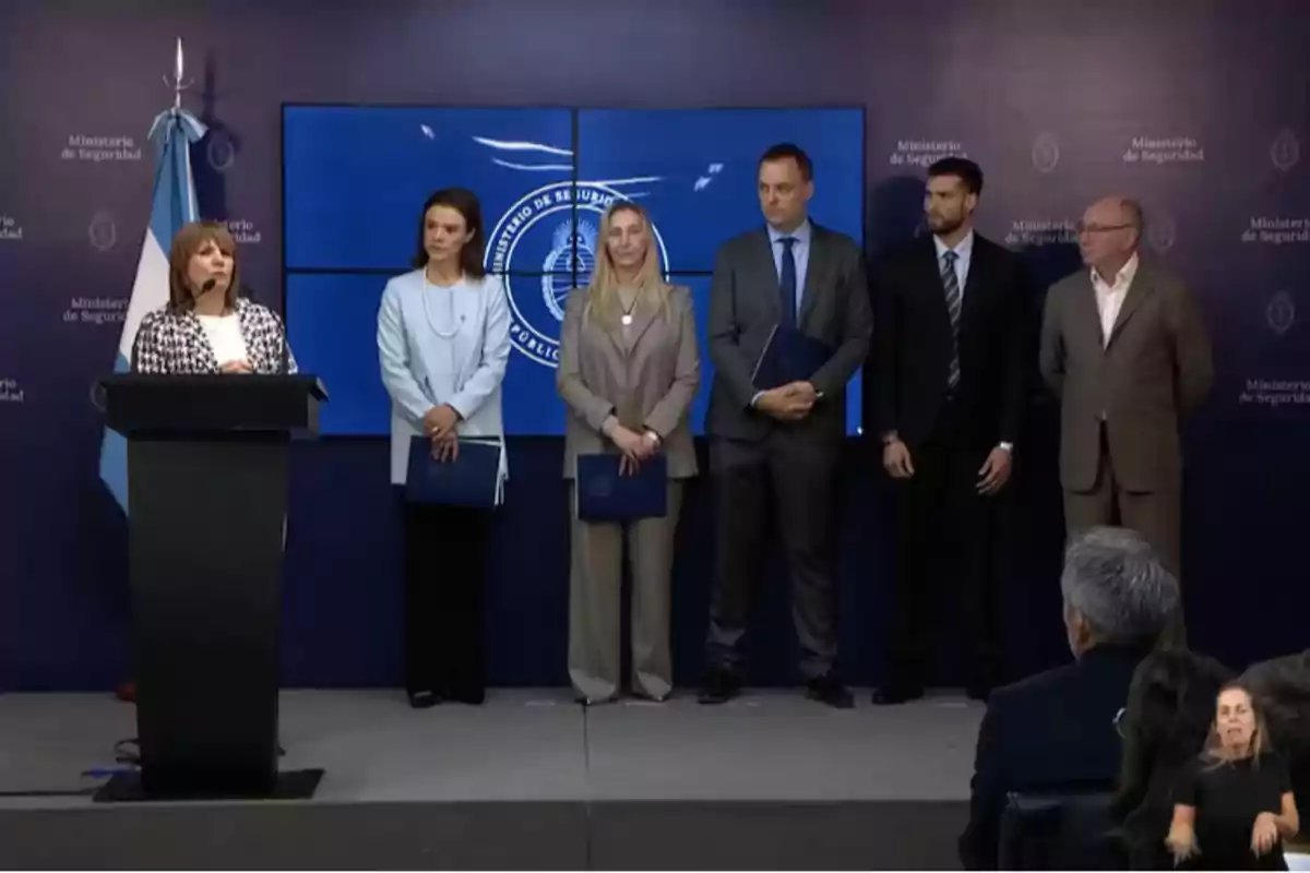 Un grupo de personas en un escenario durante una conferencia de prensa en el Ministerio de Seguridad con una bandera argentina y un intérprete de lenguaje de señas al frente. Un grupo de personas en un escenario durante una conferencia de prensa en el Ministerio de Seguridad con una bandera argentina y un intérprete de lenguaje de señas al frente.