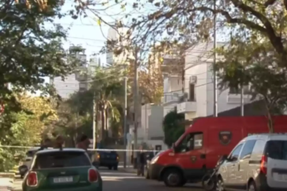 Una calle con vehículos estacionados, incluyendo una camioneta roja y un auto verde, rodeada de árboles y edificios.