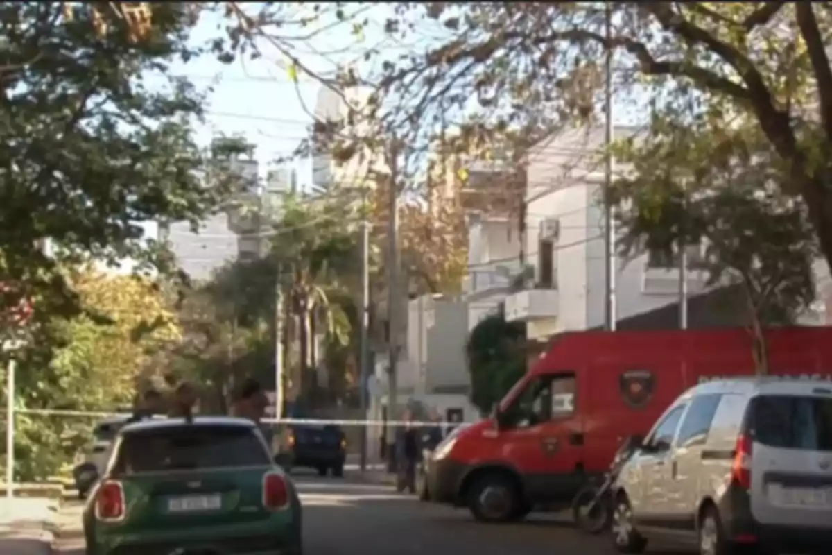 Una calle urbana con varios vehículos estacionados, incluyendo una camioneta roja y un auto verde, rodeada de árboles y edificios residenciales. Una calle urbana con varios vehículos estacionados, incluyendo una camioneta roja y un auto verde, rodeada de árboles y edificios residenciales.