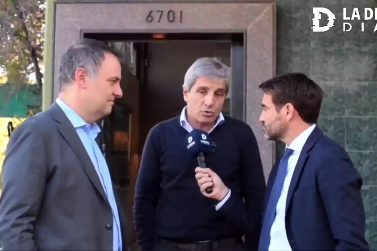 Tres hombres de pie conversan frente a una entrada numerada, uno de ellos sostiene un micrófono. Tres hombres de pie conversan frente a una entrada numerada, uno de ellos sostiene un micrófono.