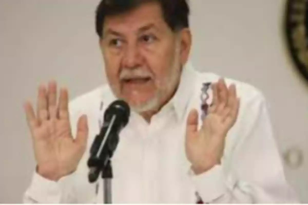 Un hombre con barba y camisa blanca habla frente a un micrófono con las manos levantadas.
