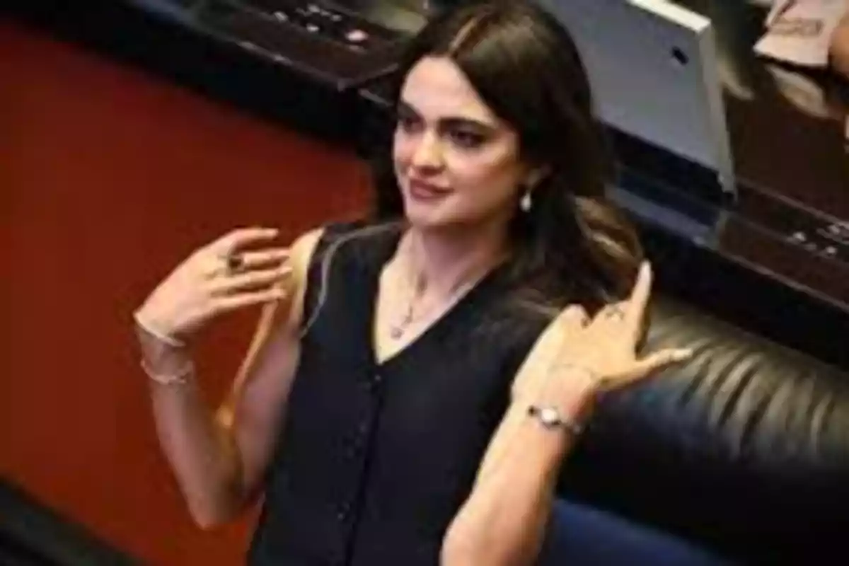 Una persona con cabello oscuro y vestida de negro está sentada en un lugar que parece ser una oficina o sala de reuniones.