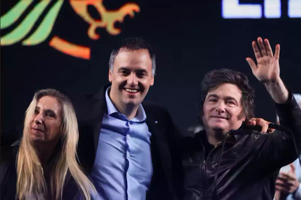 Three people smile and wave at an event, with a dark background and an illuminated logo.