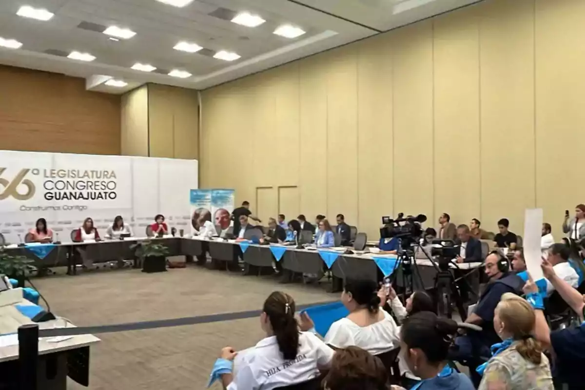 Una reunión en una sala de conferencias con personas sentadas alrededor de mesas, cámaras de video y un cartel que dice Una reunión en una sala de conferencias con personas sentadas alrededor de mesas, cámaras de video y un cartel que dice