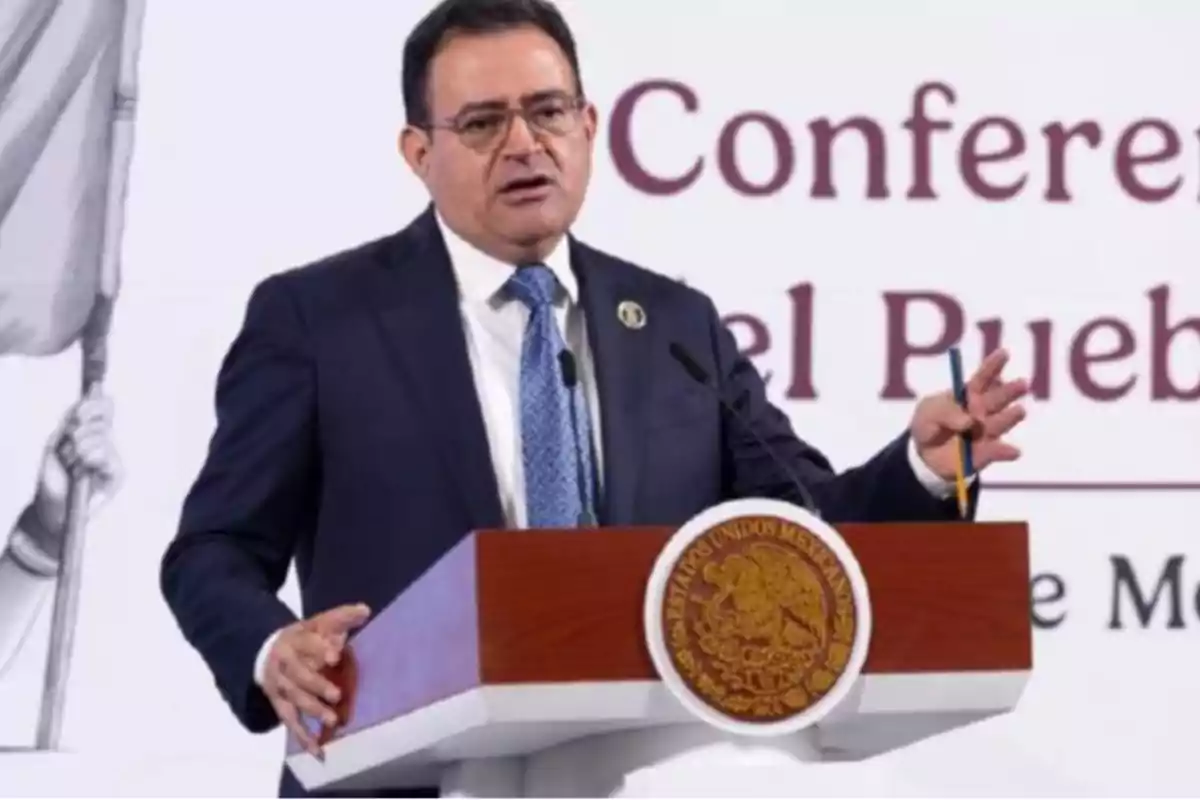 A man in a dark suit and blue tie speaks at a podium with the Mexican coat of arms.