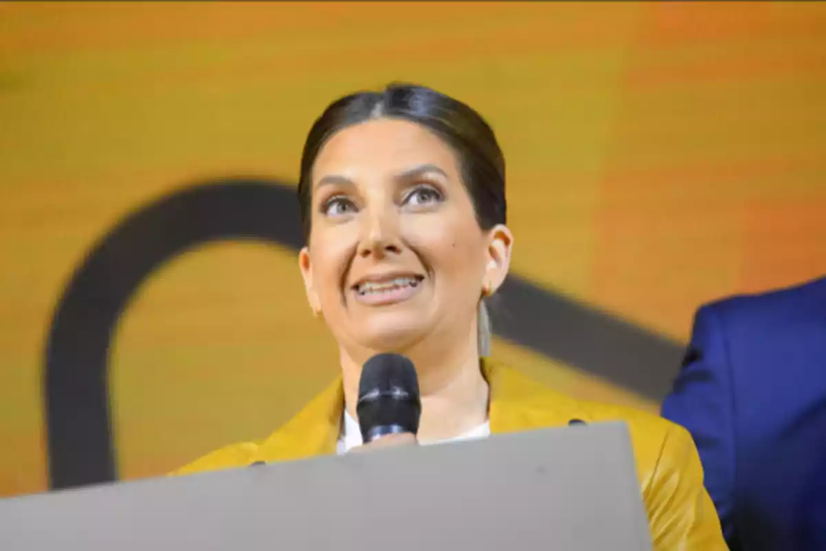 A person speaking in public with a microphone in hand and a yellow background.