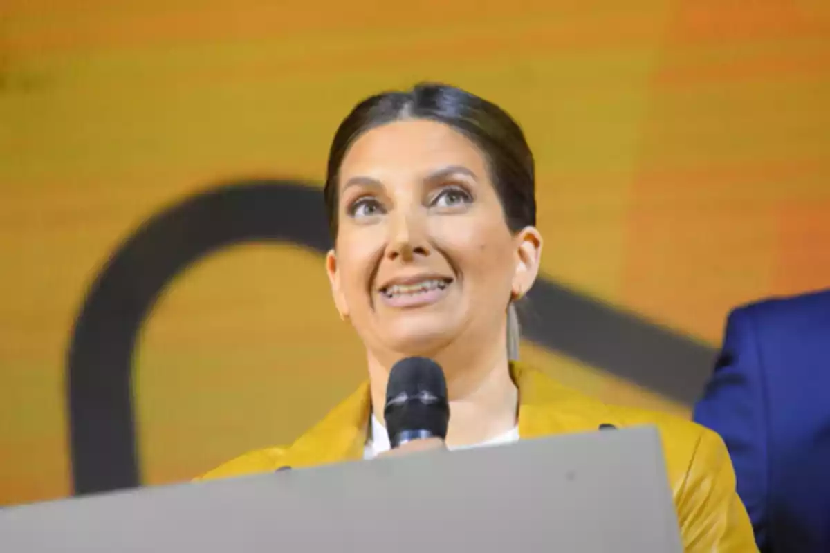 Una persona hablando en un escenario con un micrófono en la mano y un fondo amarillo.