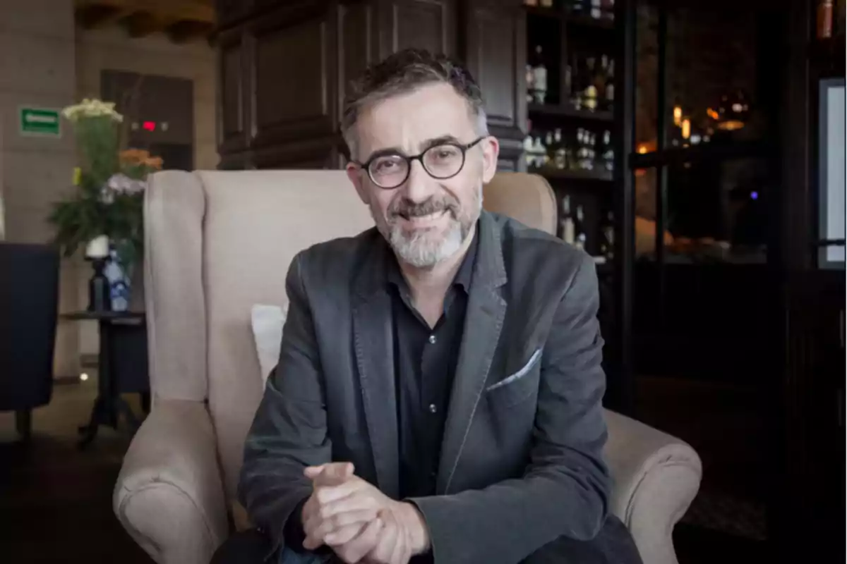Un hombre con gafas y barba sonríe mientras está sentado en un sillón en un ambiente interior elegante.