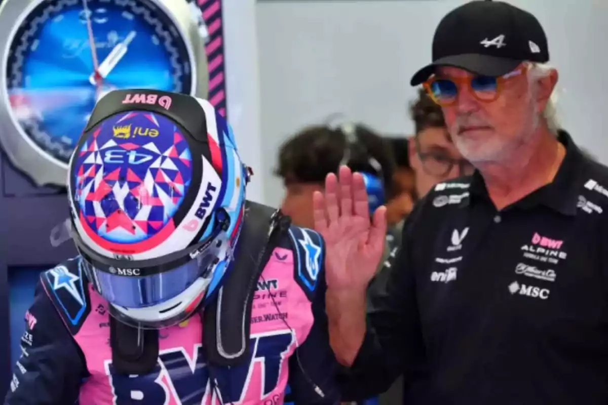 A race car driver with a pink and blue helmet and uniform is next to a man with sunglasses and a cap in a pit area. A race car driver with a pink and blue helmet and uniform is next to a man with sunglasses and a cap in a pit area.