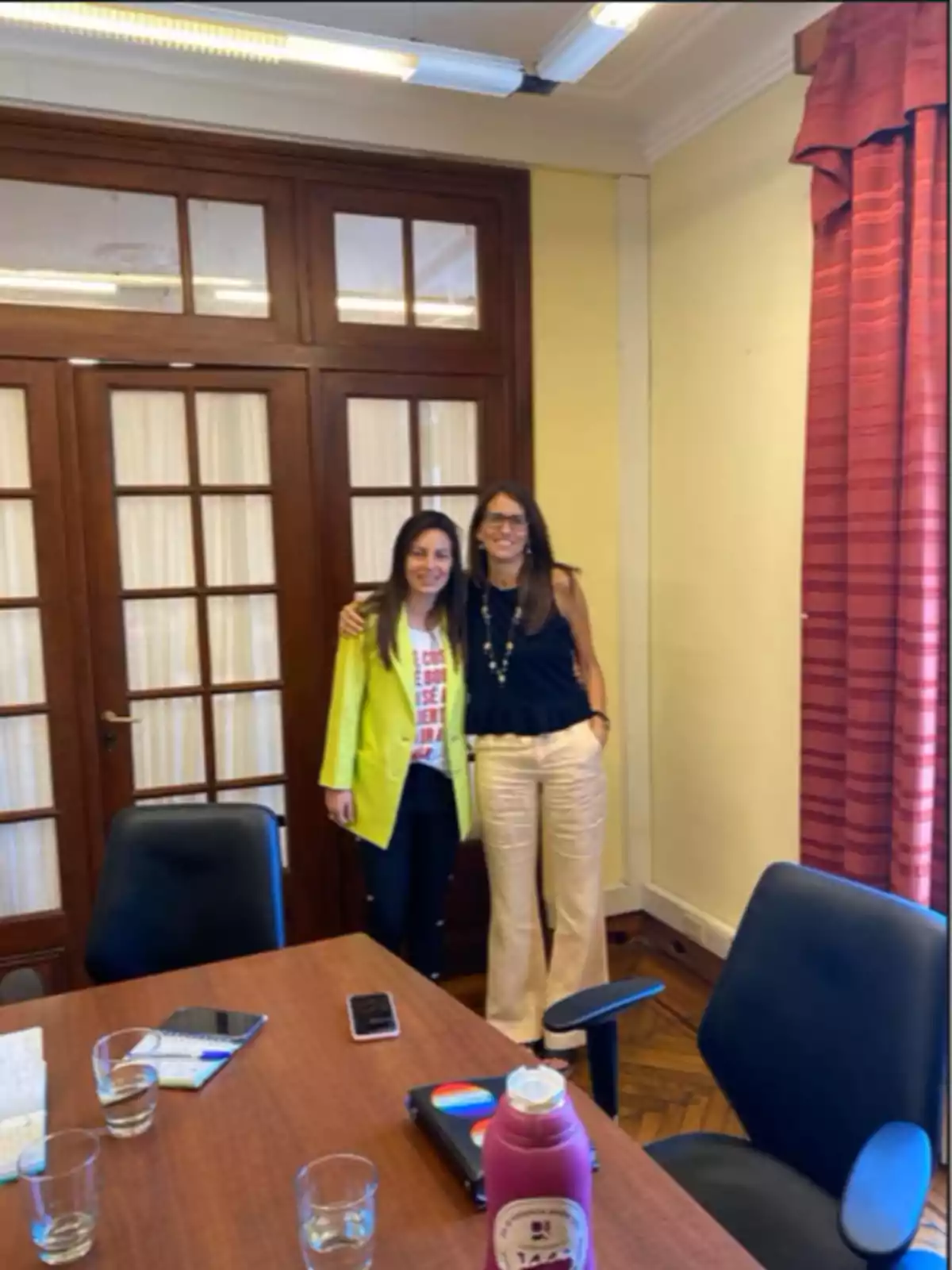 Dos personas posan juntas en una oficina con una mesa de madera, sillas y una puerta de vidrio al fondo. Dos personas posan juntas en una oficina con una mesa de madera, sillas y una puerta de vidrio al fondo.