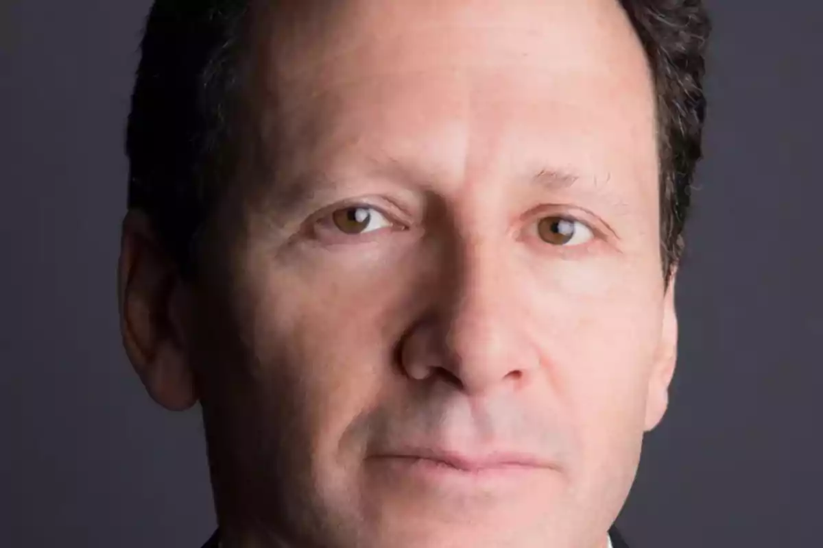A dark-haired man with a serious expression looking at the camera with a gray background.