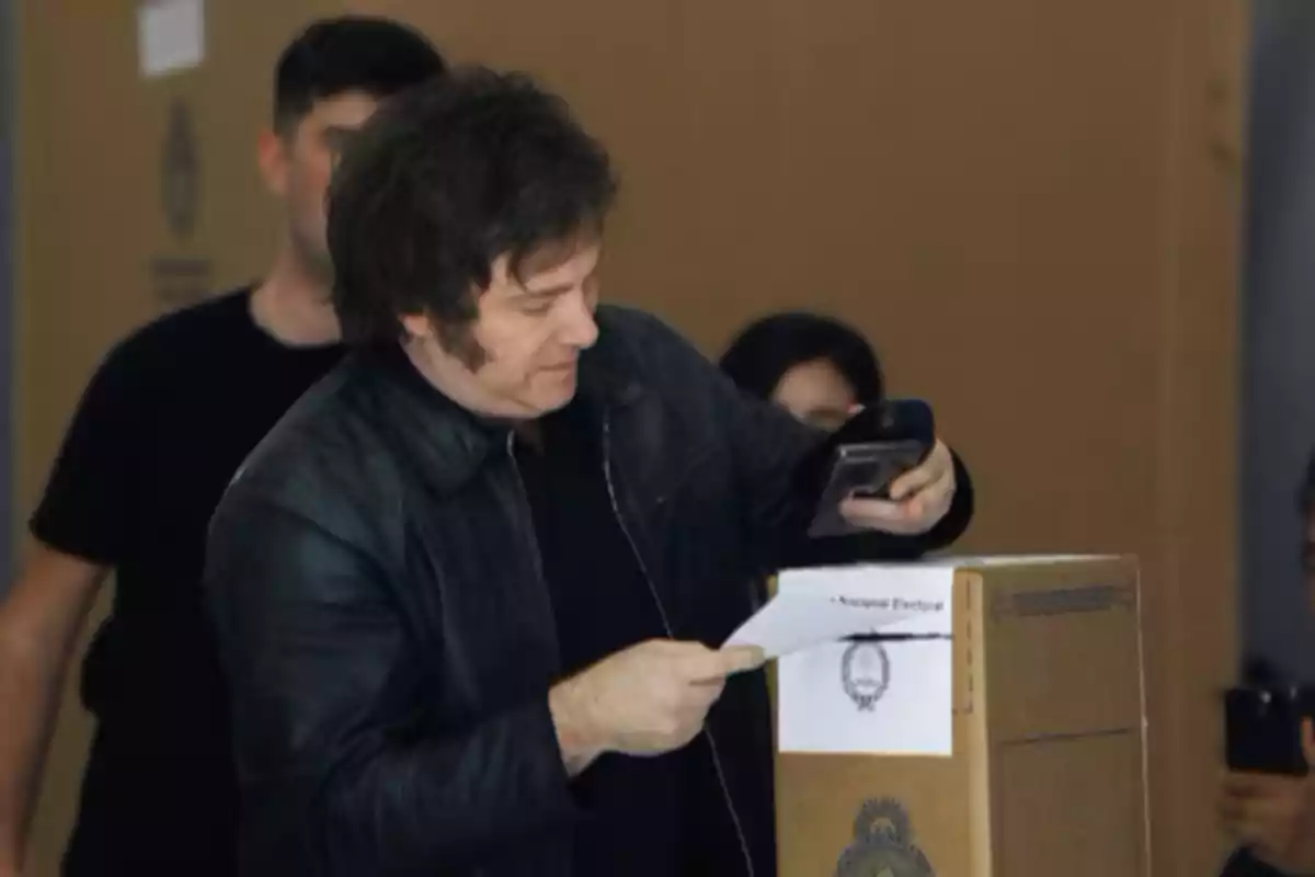 A man in a dark jacket is voting at a ballot box while holding a mobile phone.
