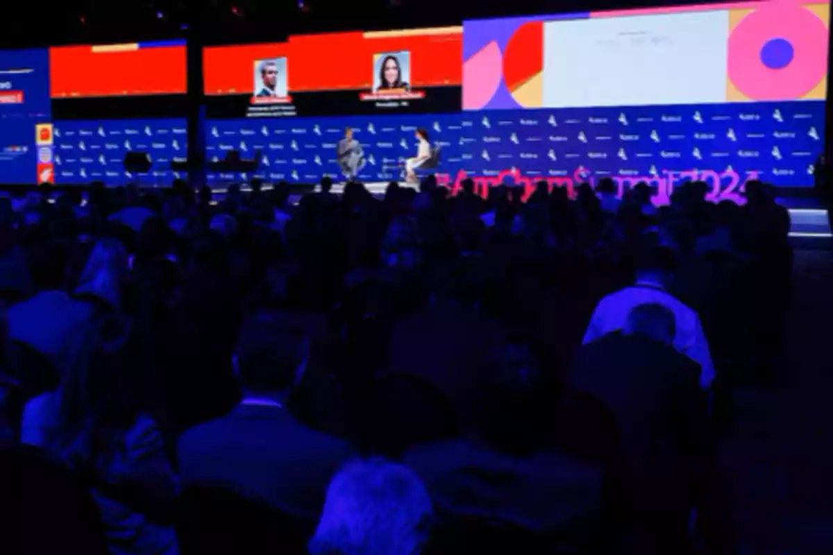 A conference with a large audience and two people on stage, with big screens displaying graphics and text. A conference with a large audience and two people on stage, with big screens displaying graphics and text.
