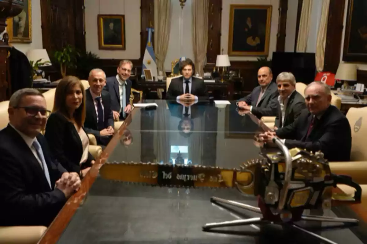 Un grupo de personas en una sala de reuniones con una bandera argentina y una motosierra sobre la mesa.