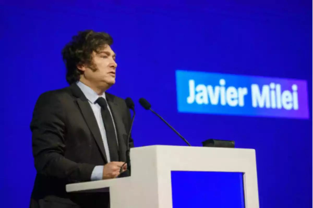 Un hombre de traje hablando en un podio con un fondo azul.
