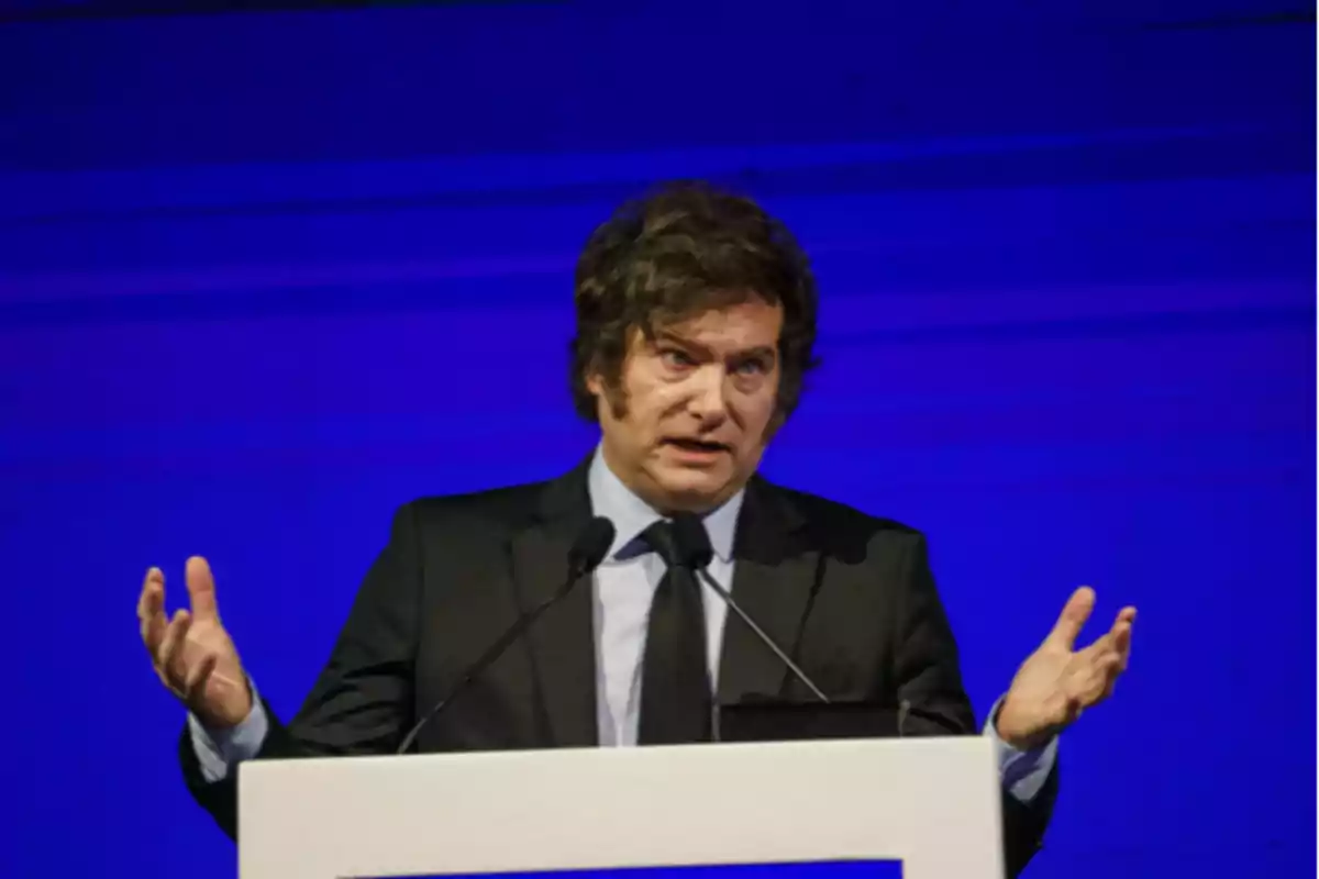 Un hombre de cabello oscuro y traje oscuro habla desde un podio con fondo azul. Un hombre de cabello oscuro y traje oscuro habla desde un podio con fondo azul.